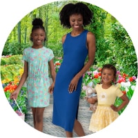 A woman and two girls in dresses stand outside, surrounded by flowers, bushes, and trees